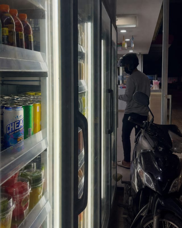 Thai convenience store at night, bright fridge doors, a motorbike parked outside the glass, a rider in a helmet inside