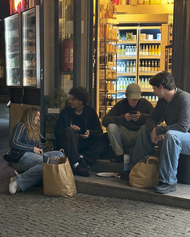 A late-night Berlin kiosk (spaeti) storefront, glowing fridge visible inside, people sitting on the step outside