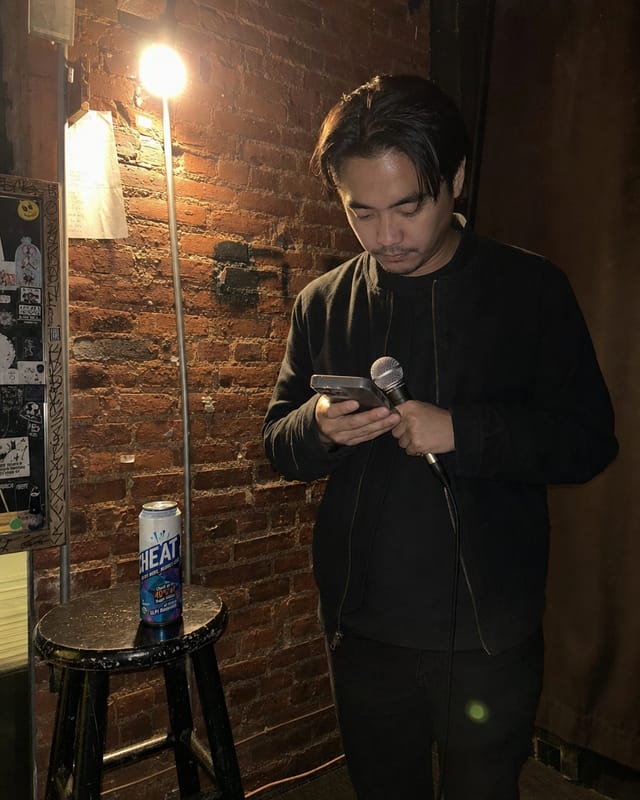 Open-mic backstage, a comedian reading notes on a phone under a bare bulb, brick wall, a stool