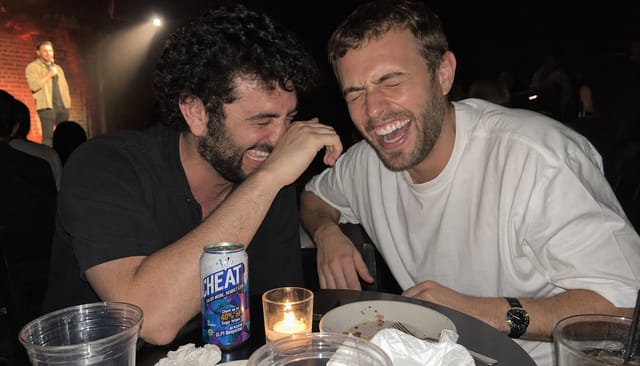 Comedy club table in the dark, a small candle, two friends crying with laughter, stage light spill