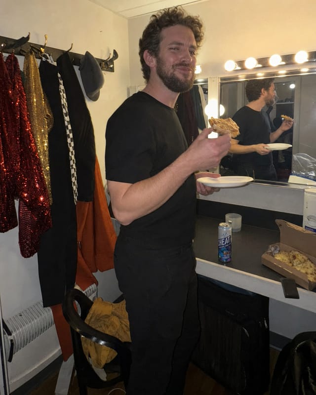 Improv troupe green room, costumes on hooks, a mirror with bulbs, someone eating a slice standing up