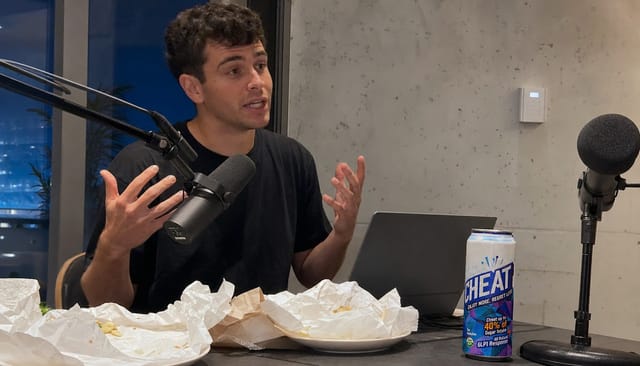 Podcast desk with two mics and a laptop, a host mid-sentence with hands up, takeaway wrappers on the desk