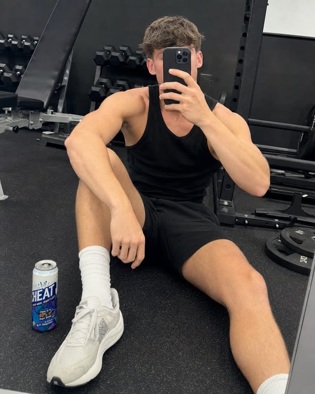 A fitness creator taking a mirror selfie in a gym, phone covering face, bright