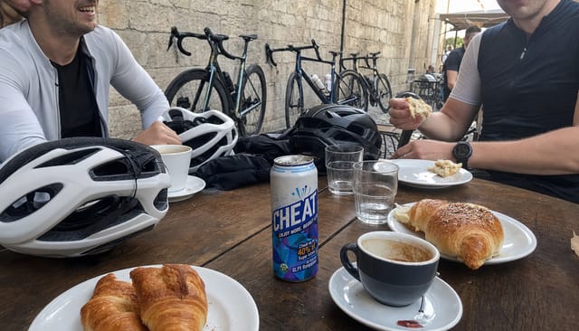 Cycling group at a cafe stop, road bikes leaning on a wall, helmets on the table, croissants