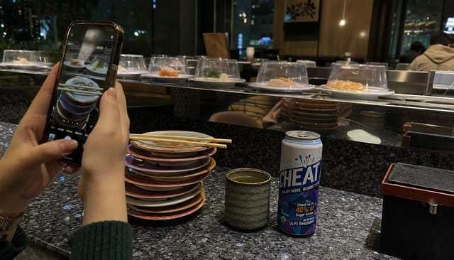 A conveyor belt sushi counter at night, plates stacked, green tea, phone photo