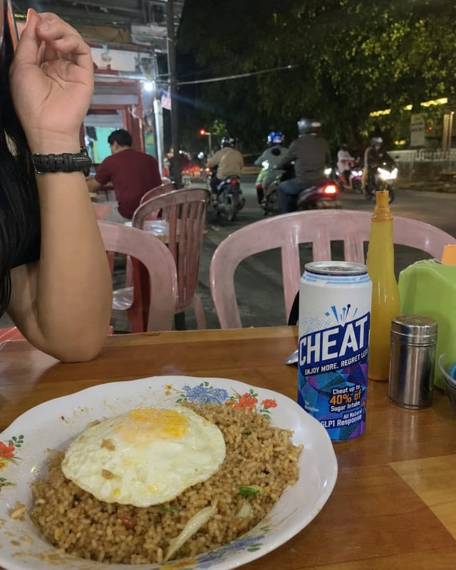 Jakarta street warung at night, a plate of nasi goreng with a fried egg, plastic chairs, motorbikes passing
