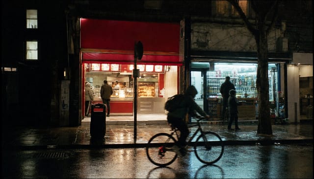 London street at night, chicken shop and off-licence fronts glowing, wet pavement, a cyclist passing