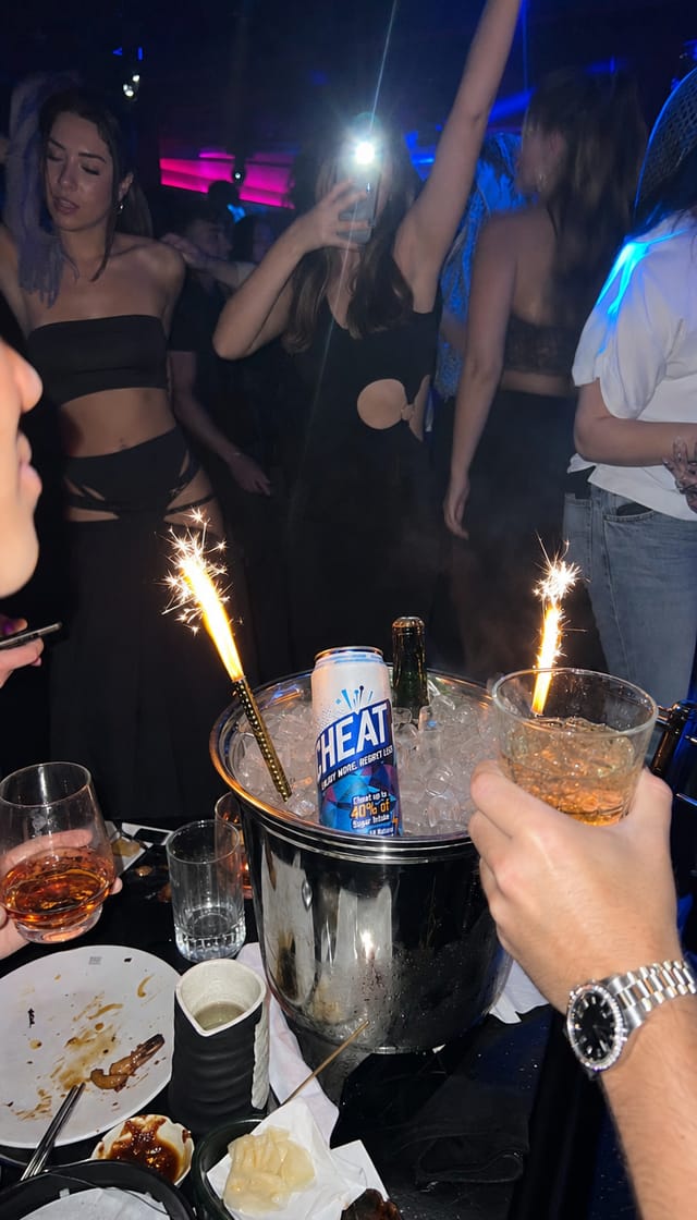 Nightclub VIP table in Miami, ice bucket with sparklers, neon pink and blue light, friends dancing in the background, flash