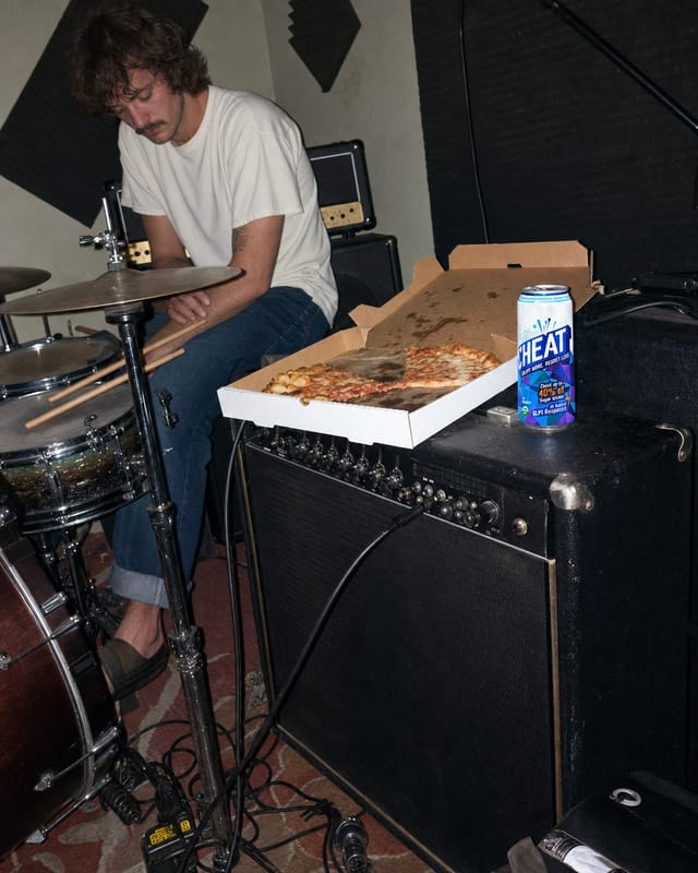 Band rehearsal room, amps, cables everywhere, a drummer resting sticks, pizza box on the amp, harsh flash