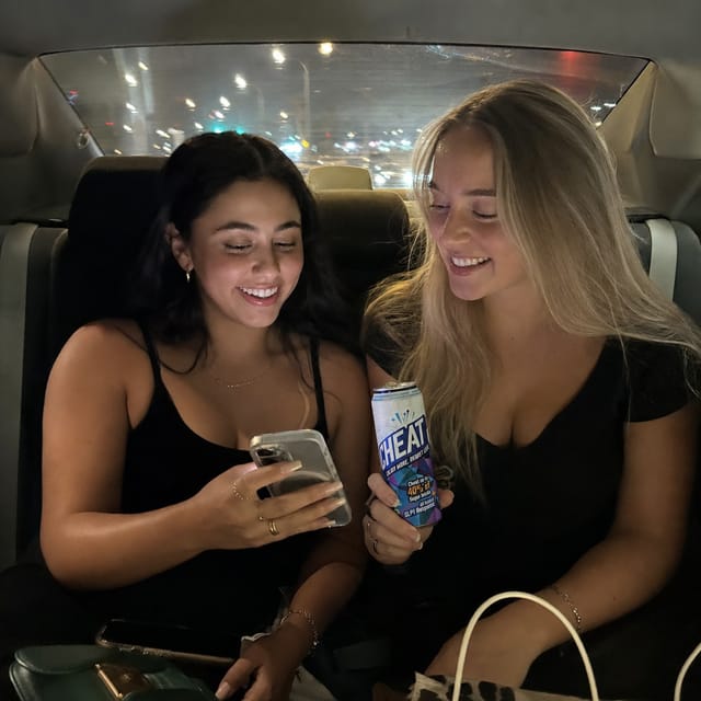 Back seat of a rideshare at night, two friends, phone light on faces, city lights streaking