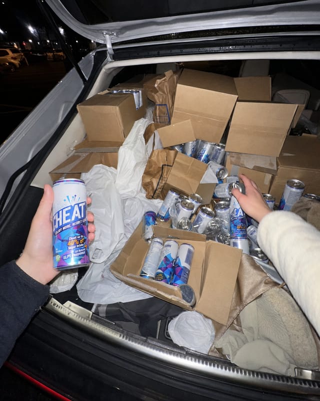 Open car boot in a dark supermarket car park packed with plain brown cardboard cases and loose cans, shopping bags squashed in, flash
