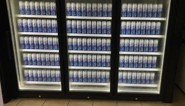 Straight-on phone photo of a three-door glass beverage fridge in a small convenience store at night, all three doors closed, the glass sligh
