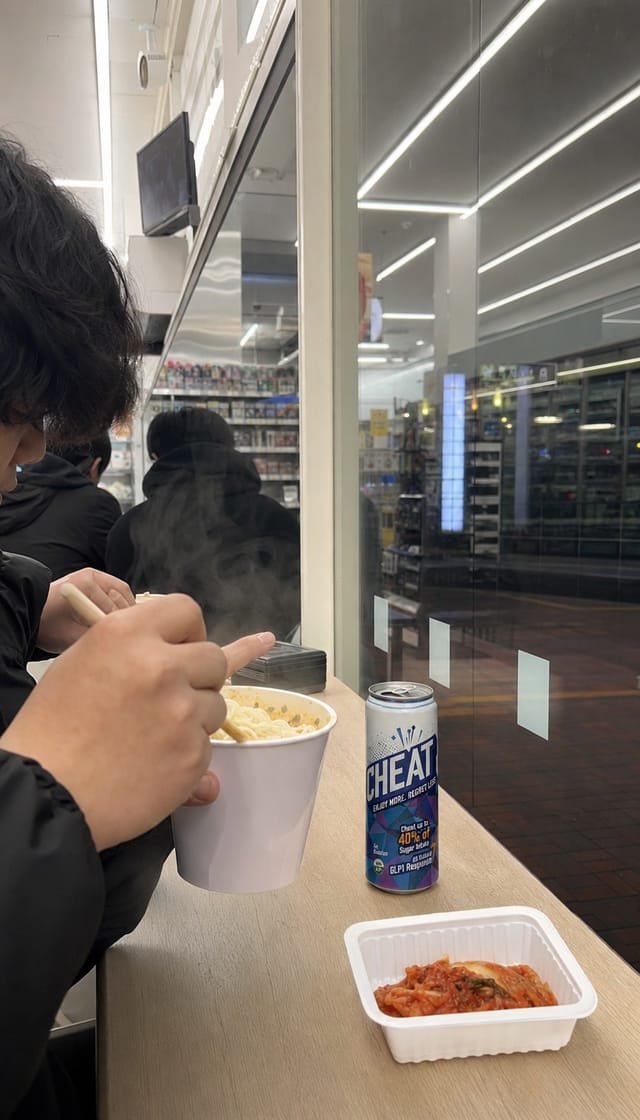 Korean convenience store interior late at night, bright white light, a small table by the window where people eat instant noodles, glass sto