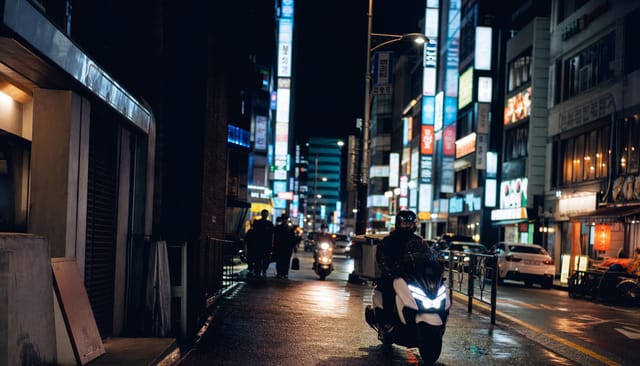 Seoul night street with dense neon signboards stacked vertically, food delivery motorbikes passing, wet asphalt