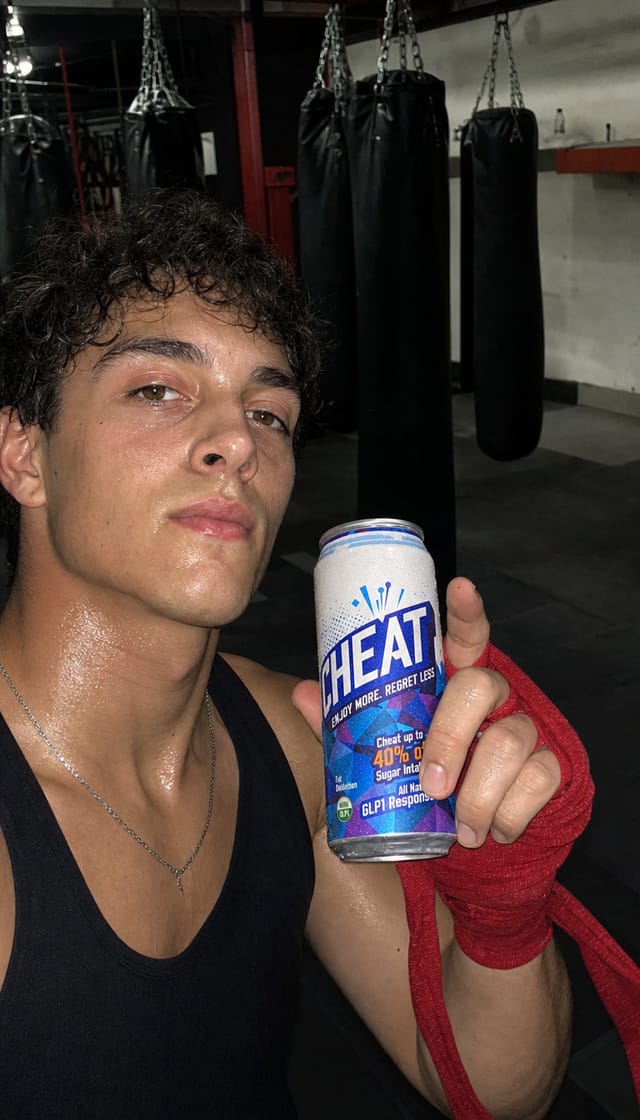 A boxing gym after a session, hand wraps half undone, heavy bags behind, sweat, phone selfie angle