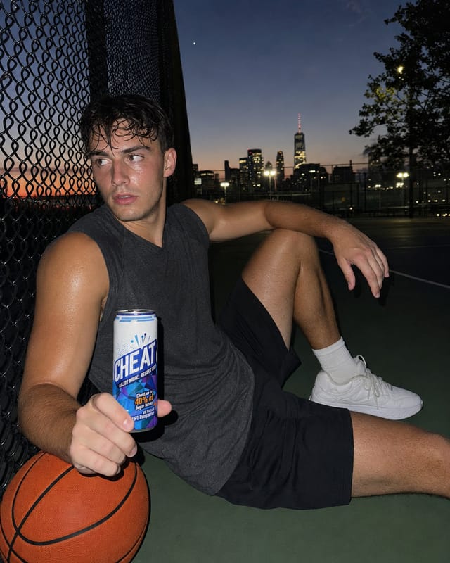 Outdoor basketball court at dusk, a sweaty player sitting against the fence taking a break, ball on the ground, city behind, flash