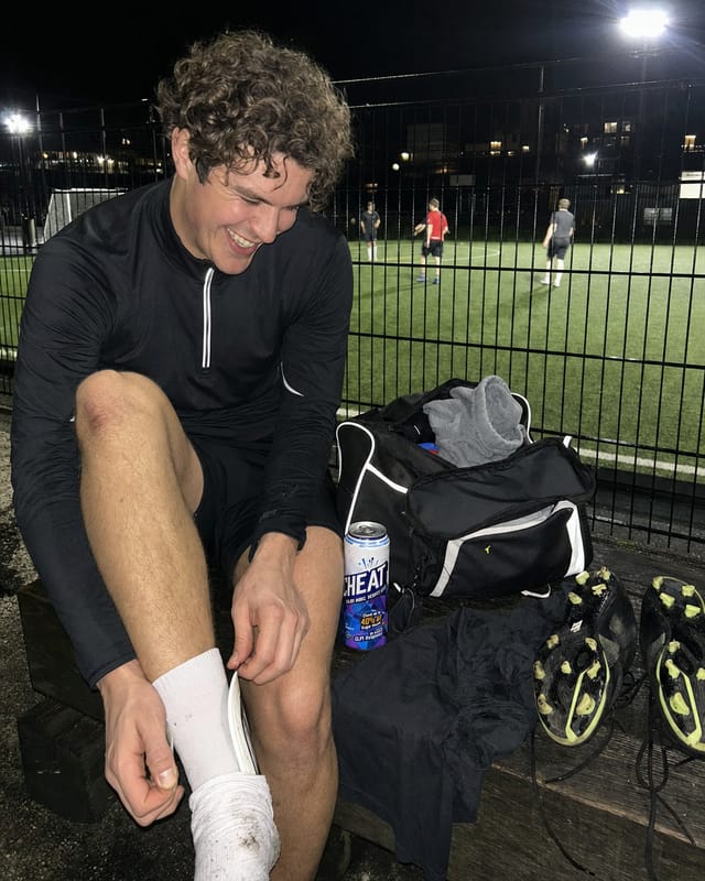 Sideline of a five-a-side football pitch at night under floodlights, kit bags and boots, a player mid-laugh pulling off a shin pad
