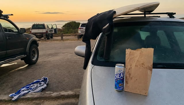 Surf car park at dawn, a wetsuit half peeled, board on the roof rack, towel on the ground, takeaway bag