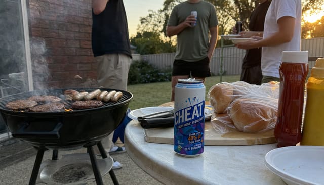 Backyard BBQ with a smoking grill, sausages and burgers, plastic table with buns and sauces, friends in the background, golden hour