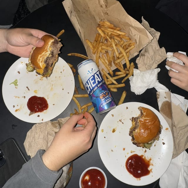 Overhead shot of a messy table with two smash burgers, fries spilled from a paper bag, ketchup, crumpled napkins, flash