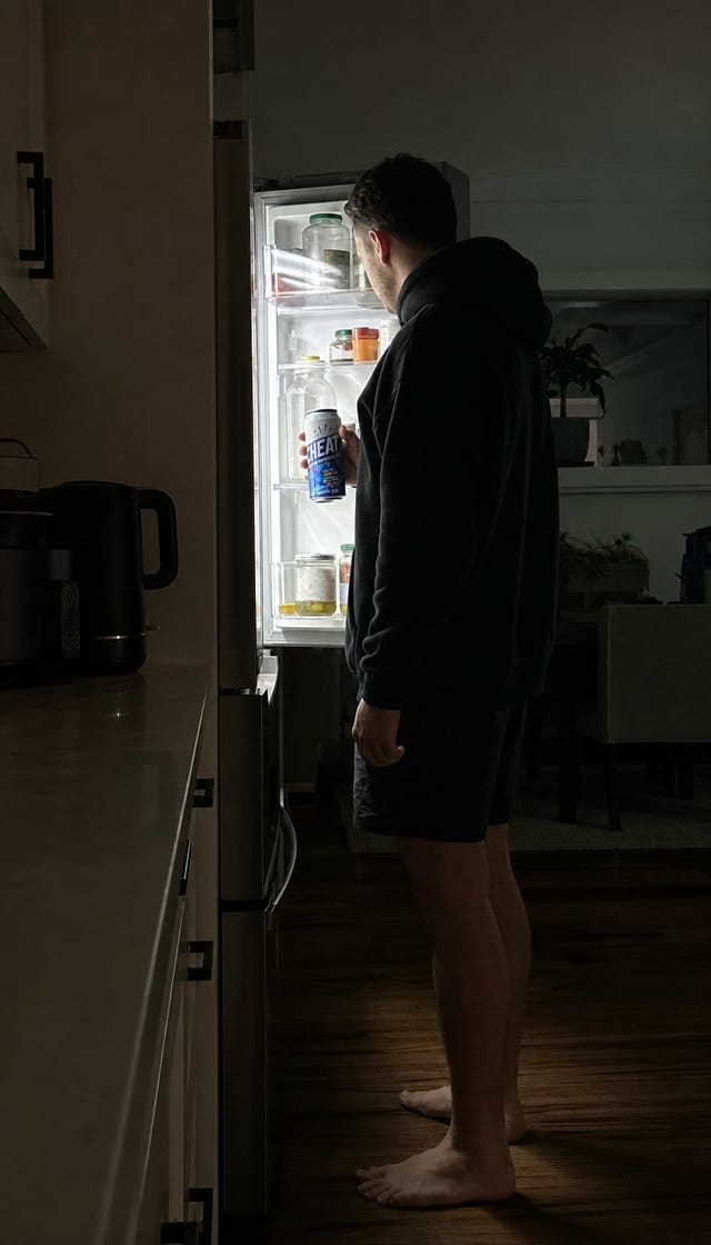 A person standing in front of an open fridge at night in a dark kitchen, lit only by the fridge light, bare feet, hoodie