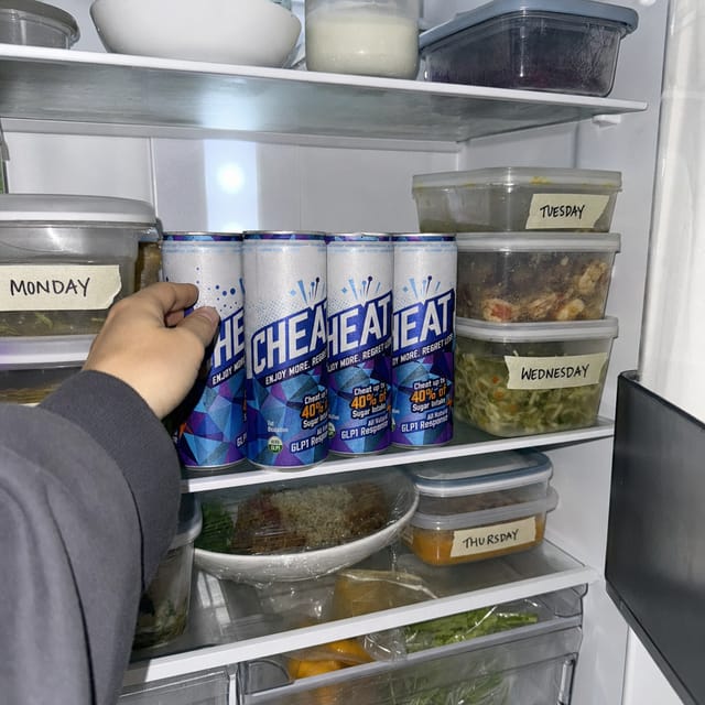 Open office kitchen fridge with labelled lunch boxes and drinks, fluorescent light, someone's hand reaching in