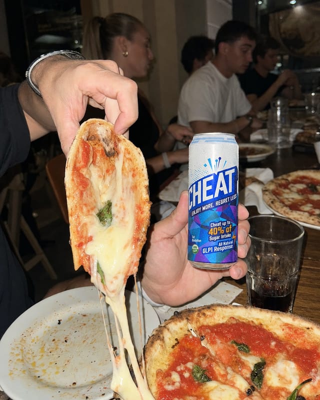 A person's hand holding a folded pizza slice toward the camera in a busy pizzeria, cheese pull, other hand reaching for a drink, flash