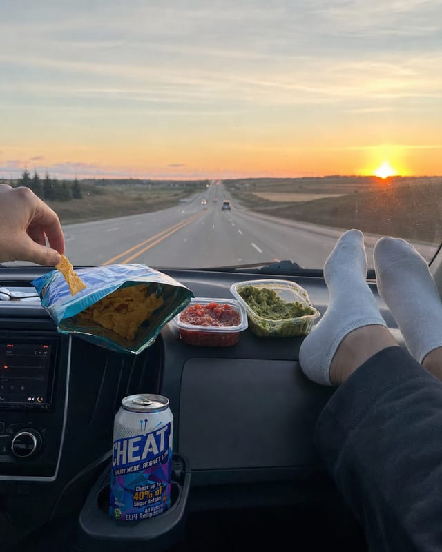 Car dashboard on a road trip at sunset, snacks on the dash, highway ahead, feet on the dash